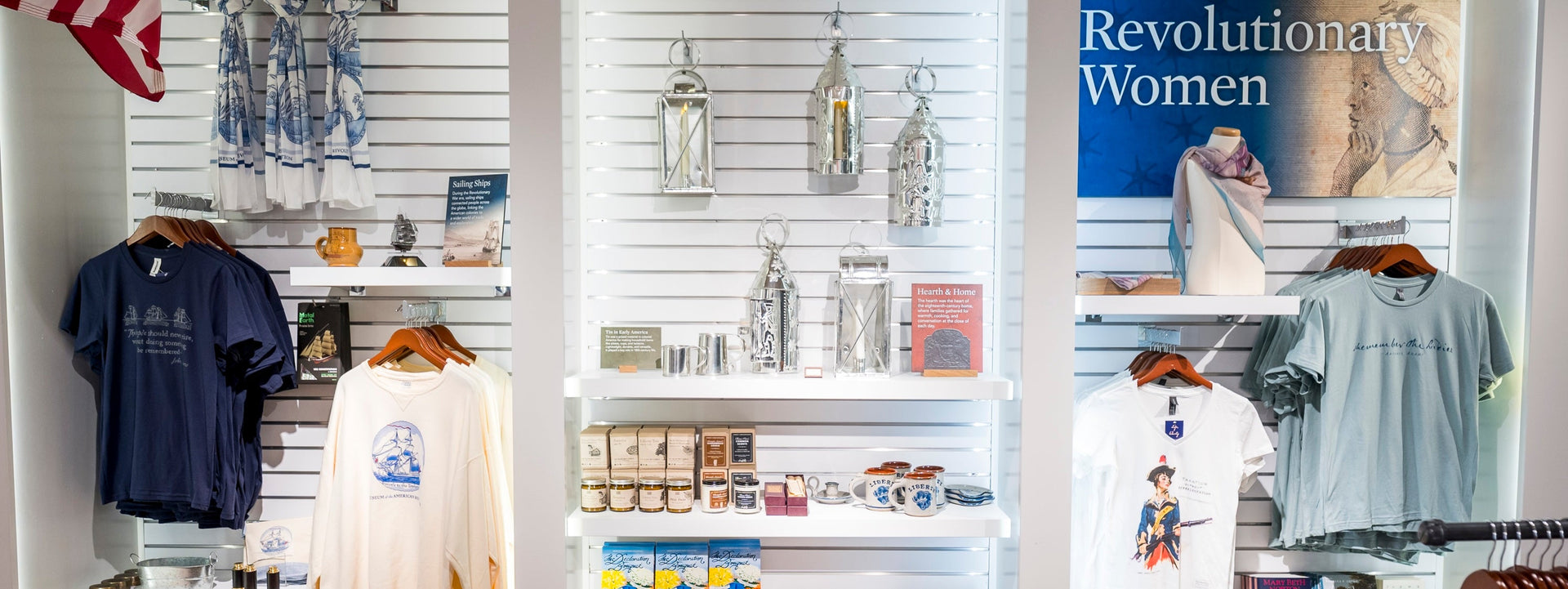 Gift shop display featuring historical-themed T-shirts, mugs, books, lanterns, and souvenirs, with a banner on the right reading “Revolutionary Women” and part of an American flag visible on the left.
