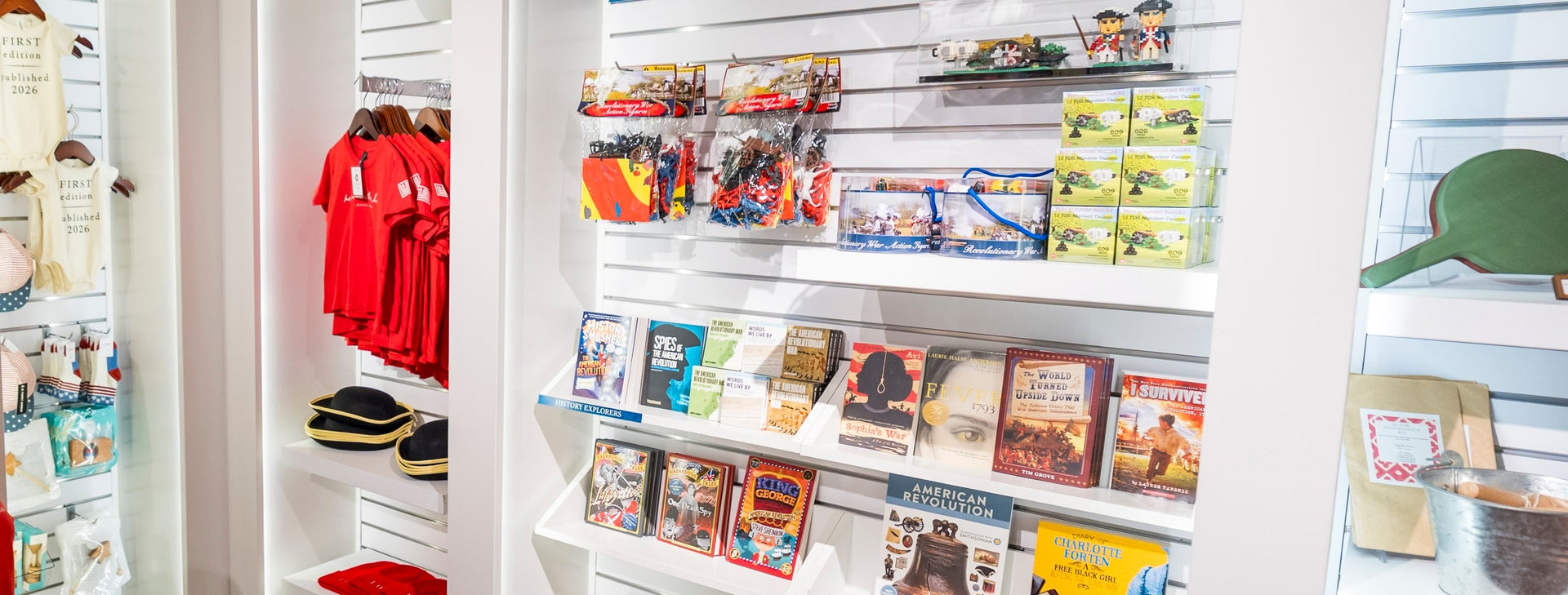 Gift shop display with red t-shirts, toy soldier hats, Playmobil sets, puzzles, model kits, and history books on white shelves. Items have a historical and patriotic theme.