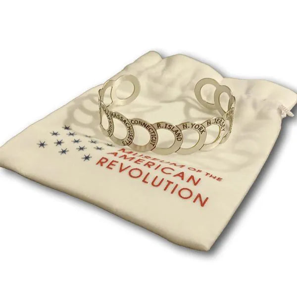 The Chain of States Bracelet, featuring silver circular cutouts engraved with state names, rests on a white pouch labeled "American Revolution" and decorated with blue stars.