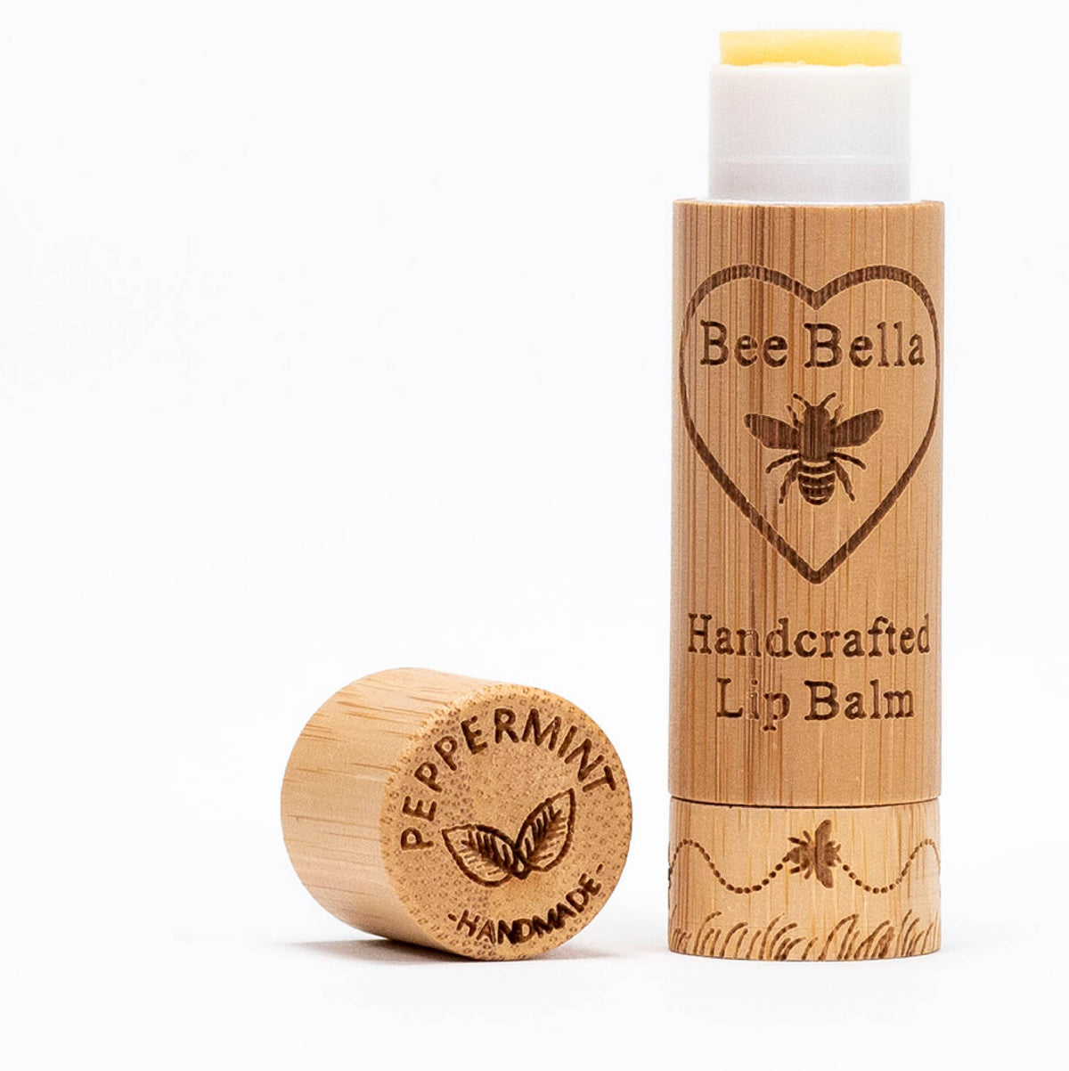 A cylindrical wooden tube of Bee Bella Peppermint Lip Balm with its cap removed, featuring engravings of a bee inside a heart, the text Bee Bella Handcrafted Lip Balm, and Peppermint Handmade on the cap.