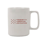 The Seneca Falls Mug is a white ceramic mug featuring the Museum of the American Revolution logo in red and a cluster of stars beside the museum name, commemorating the Women’s Rights Convention.
