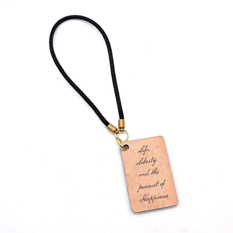 The "Life, Liberty, and the Pursuit of Happiness" Bag Charm is a rectangular tan leather keychain with a black cord and gold hardware, engraved in cursive.