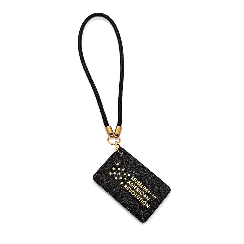 The "Life, Liberty, and the Pursuit of Happiness" Bag Charm features a black rectangle with thirteen gold stars in a circle, a gold clasp and black loop.