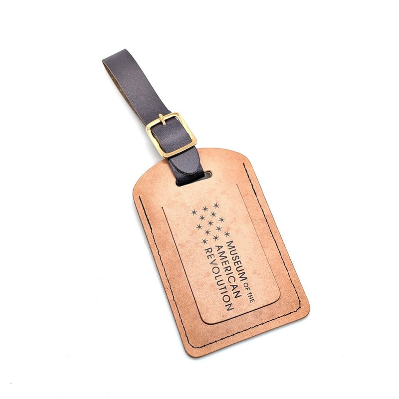 The "Life, Liberty, and the Pursuit of Happiness" tan leather luggage tag has a black buckle strap and an engraved thirteen-star design with Museum of the American Revolution text.