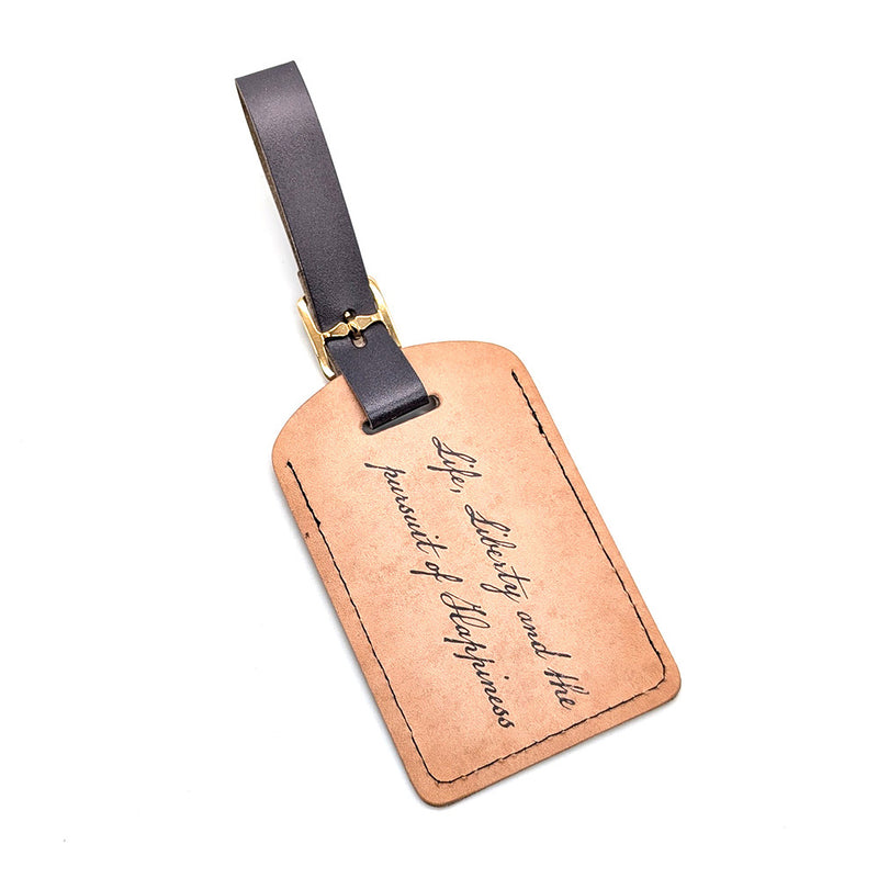 The "Life, Liberty, and the Pursuit of Happiness" Luggage Tag is crafted from durable brown leather with a black strap and gold buckle, featuring elegant cursive engraved text.