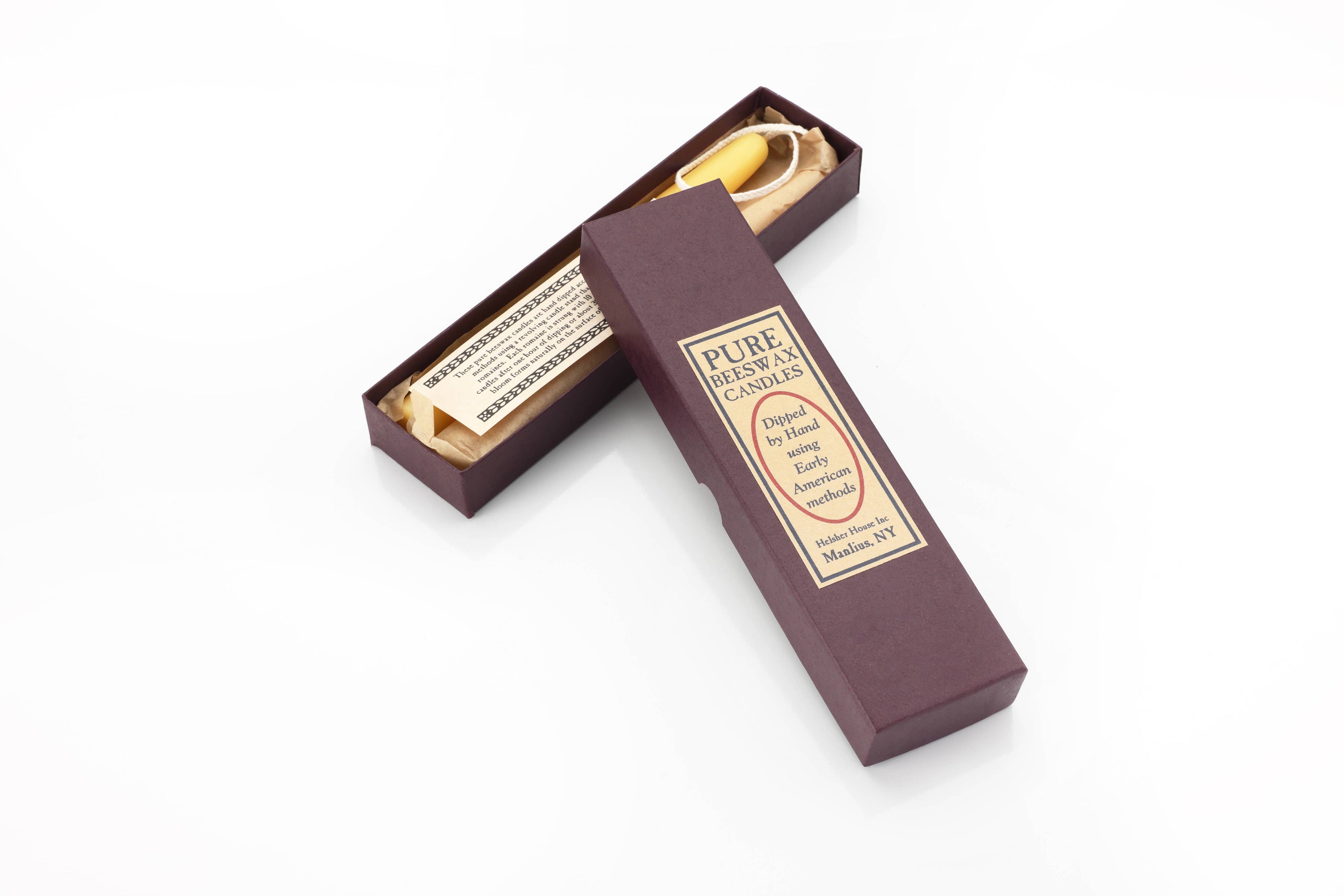 A dark purple box labeled “Beeswax Taper Candle” rests open on a white surface, displaying a hand-rolled yellow beeswax taper candle and an informational slip inside.