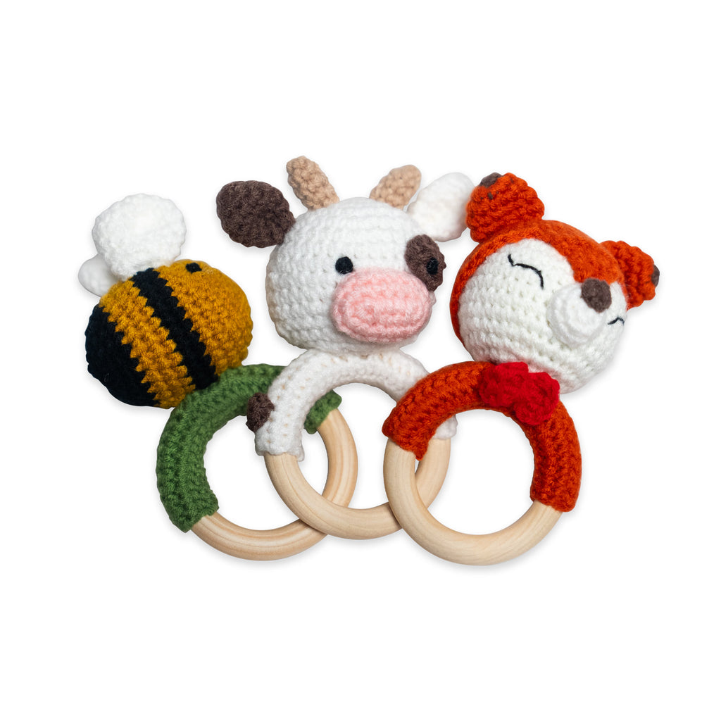 Three Hand Crochet Rattle toys, each shaped like a bee, a cow, and a fox, with wooden teething rings, are arranged overlapping on a white background. Each Hand Crochet Rattle is crafted from organic cotton for a safe and charming baby toy.