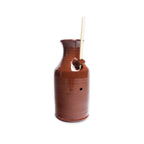 A Bird Bottle, crafted from brown ceramic and featuring a small spout with a hole near the middle that holds two thin wooden sticks, is displayed against a white background.