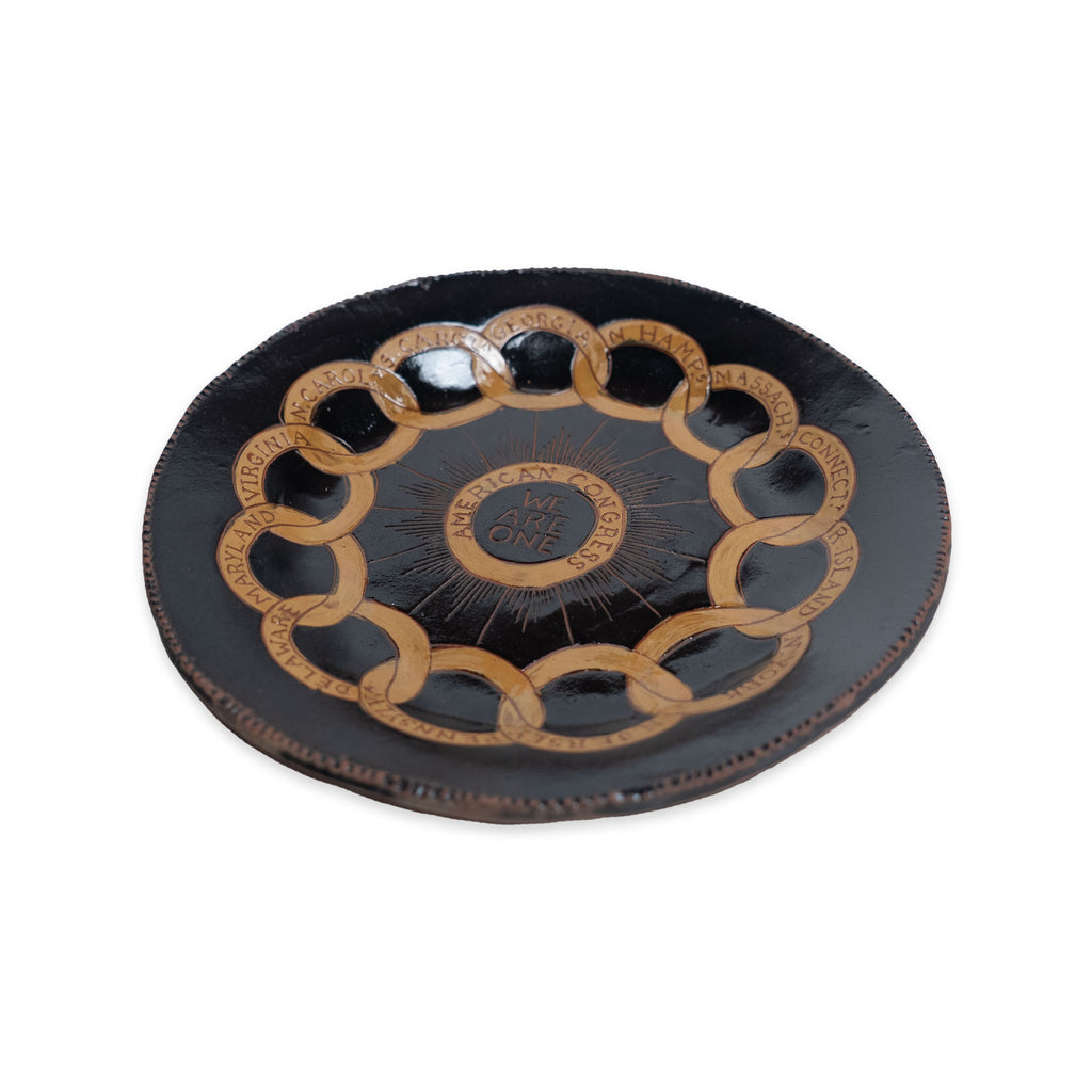The Chain of States Plate, Black is a handcrafted ceramic plate with interlocking gold rings and engraved text along the rim, featuring “We Are One” in the center.