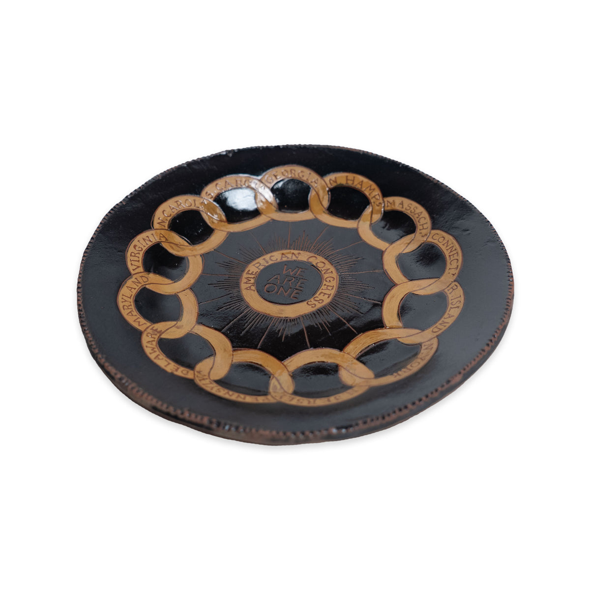The Chain of States Plate, Black is a handcrafted ceramic plate with interlocking gold rings and engraved text along the rim, featuring “We Are One” in the center.
