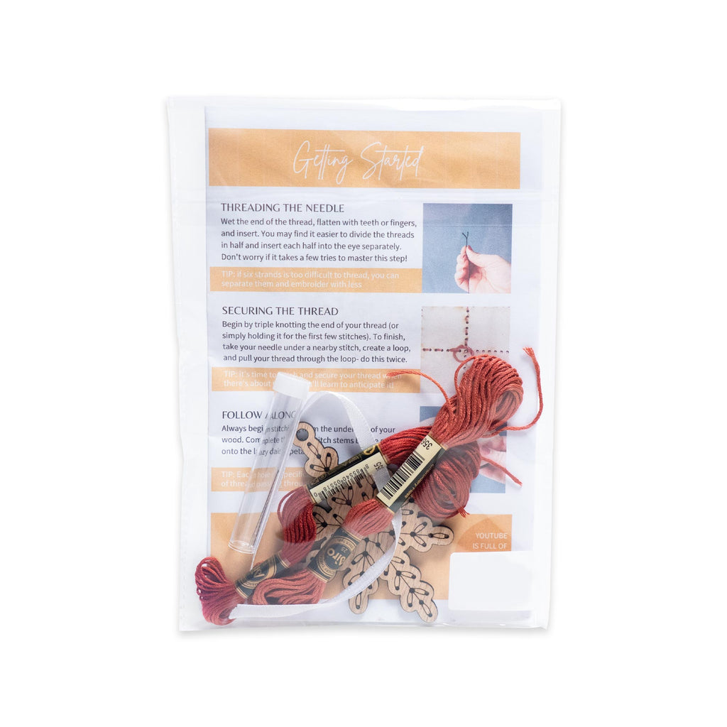 Snowflake Ornament Embroidery Kit packaged in a clear plastic sleeve with instructional cards, a wooden snowflake cutout, and a skein of dark red embroidery floss.