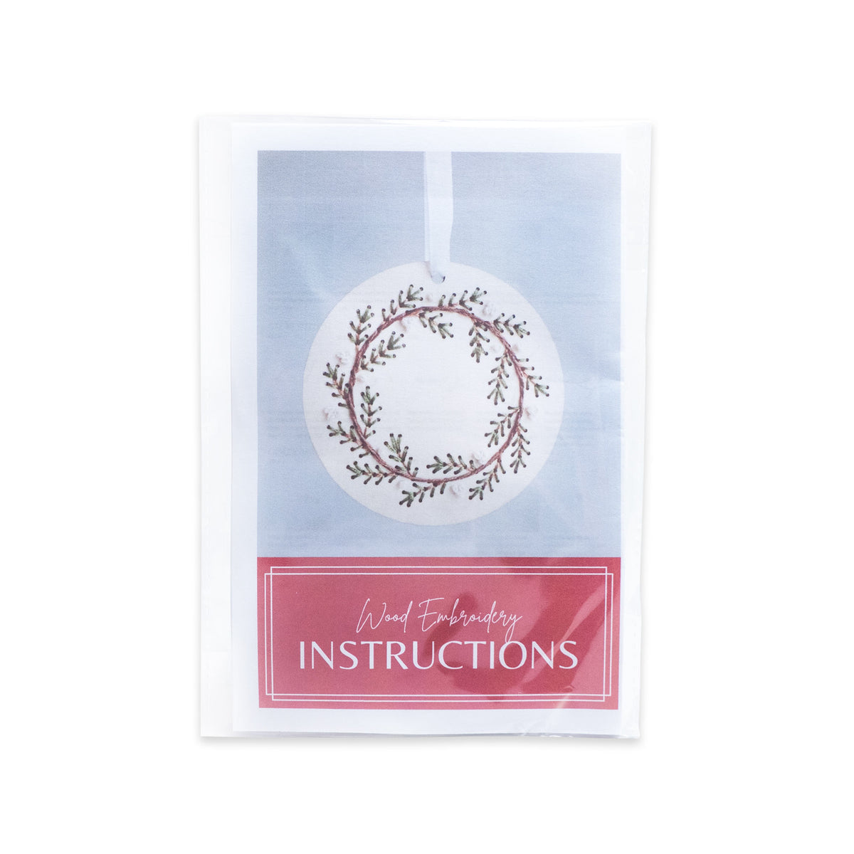Pine Wreath Ornament Embroidery Kit in a clear plastic sleeve, showing a circular wood ornament with an embroidered wreath design above the text Wood Embroidery INSTRUCTIONS.