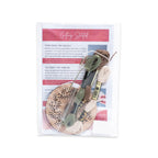 A Pine Wreath Ornament Embroidery Kit in a clear plastic bag containing a printed instruction sheet, a small wooden embroidery hoop, and three skeins of embroidery floss in shades of green, brown, and cream.