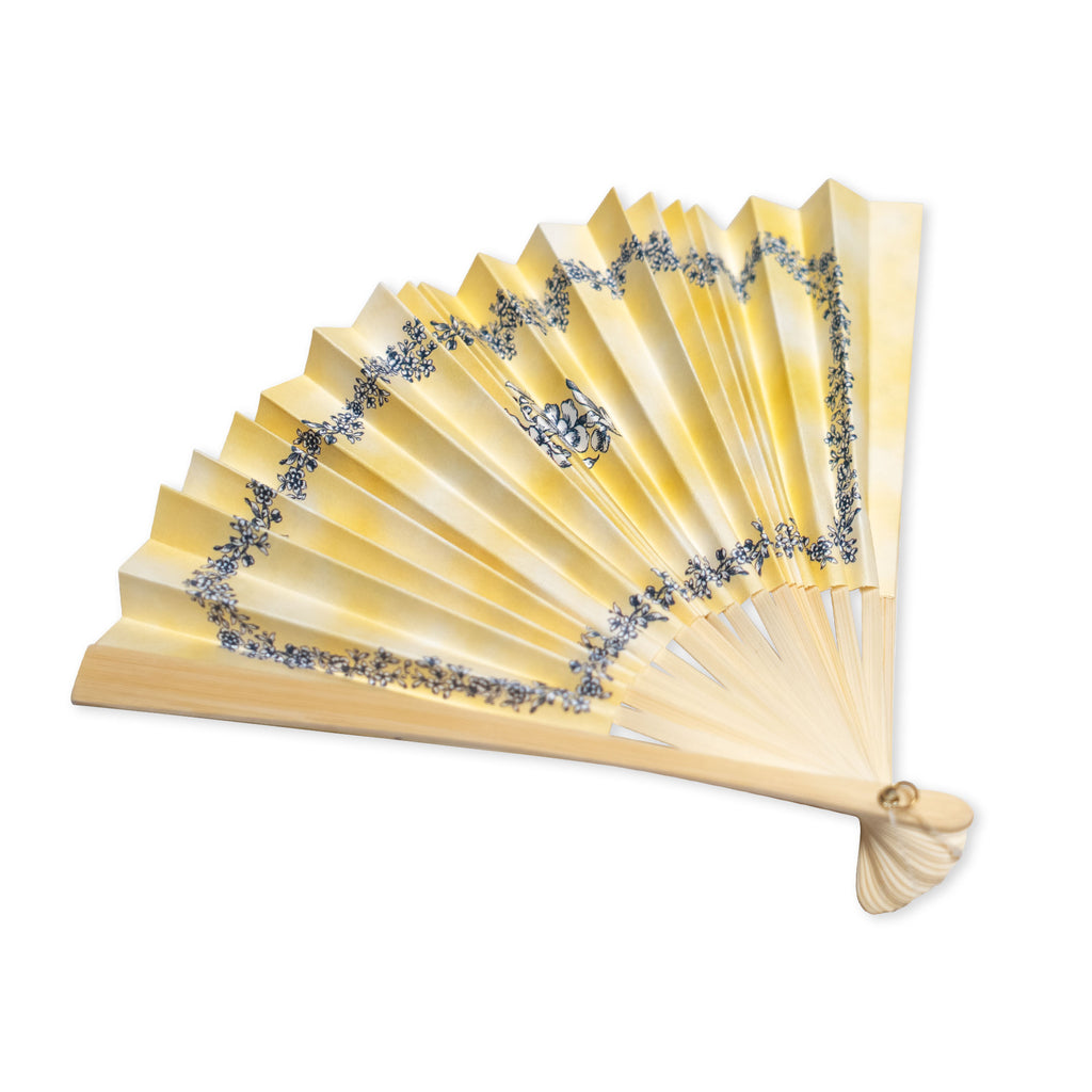 The Moral Compass Fan, a yellow folding hand fan adorned with blue floral patterns, is open and elegantly displayed on a white background, serving as a decorative accessory that evokes historical charm.