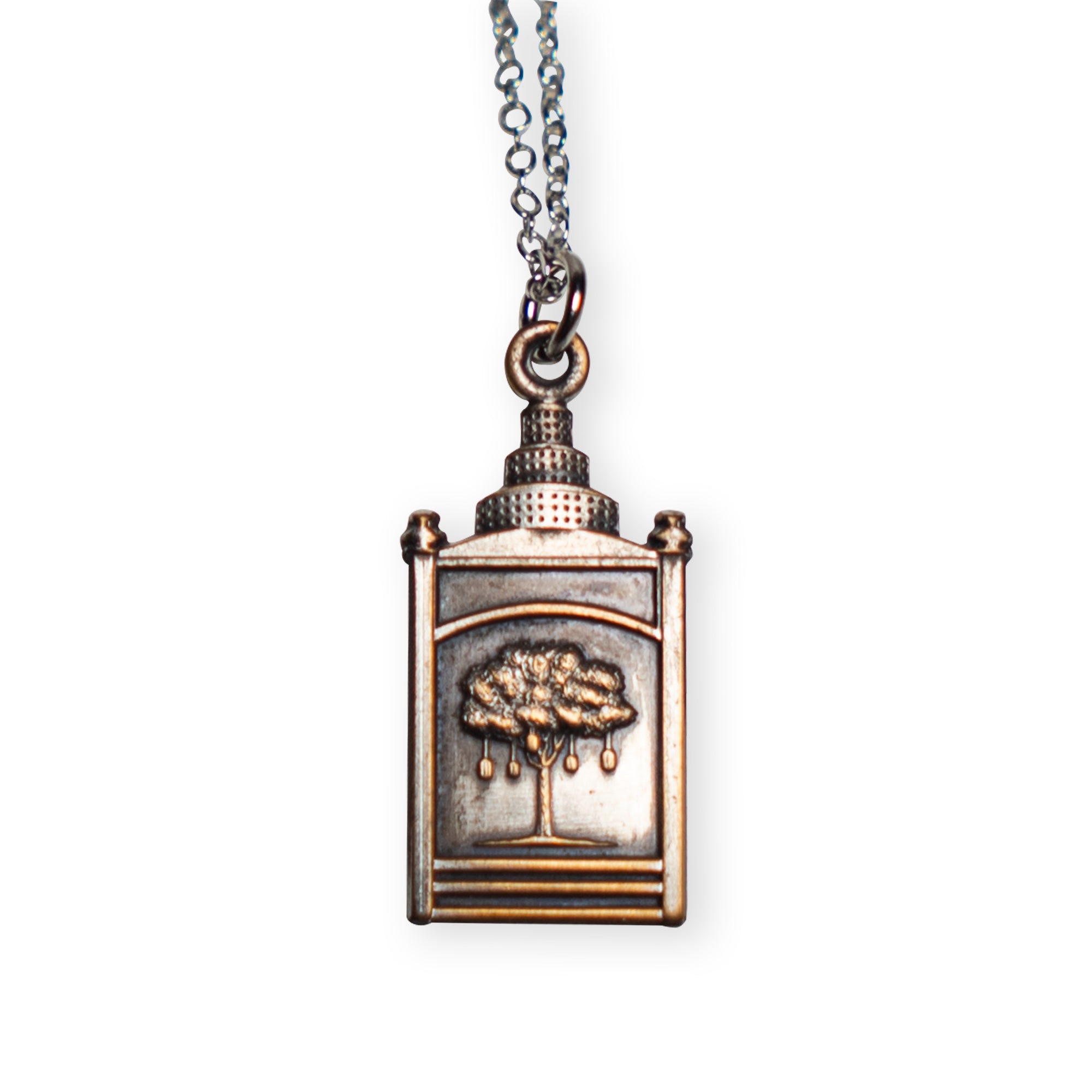 The Liberty Tree Necklace is a silver pendant shaped like a small book, featuring an embossed tree with hanging ornaments on the cover, inspired by the historic Liberty Tree. Its antique silver finish and delicate silver chain make this piece a timeless Boston symbol, beautifully displayed against a white background.