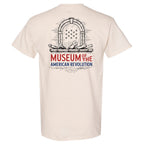 The Museum T-Shirt, Short Sleeve, is a beige tee with a sketch of an arch, cannons, and stars above "Museum of the American Revolution" in bold blue and red text on the back.
