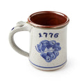 The 1776 Stoneware Mug is handcrafted with a beige exterior, blue floral design, and "1776" in blue. Its interior features a rich reddish-brown glaze.