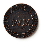 A close-up of a 1776 New Hampshire Halfpenny, an old colonial coin featuring large WM letters at its center and worn raised text around the edge that partially reads "AMERICAN LIBERTY.