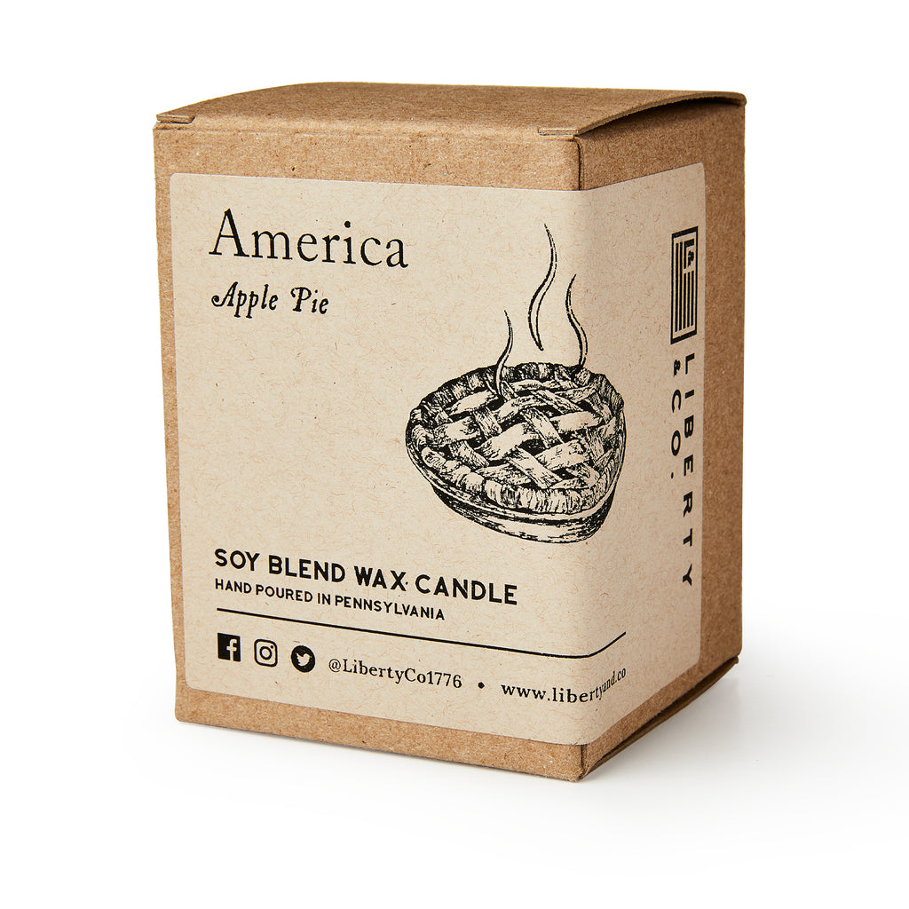 A brown cardboard box labeled America Candle holds a soy blend candle. The label shows a steaming apple pie and notes it's a hand-poured apple pie candle, perfect for making your home cozy.