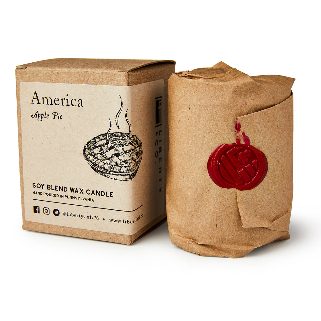 A boxed America Candle with an apple pie illustration sits next to a brown paper-wrapped candle sealed with red wax—perfect for adding a warm, inviting apple pie aroma to any space.