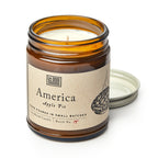 An America Candle in a brown glass jar, labeled "America Apple Pie" with a pie drawing and flag icon, sits with its metal lid. The partially used white soy blend candle creates a cozy atmosphere.