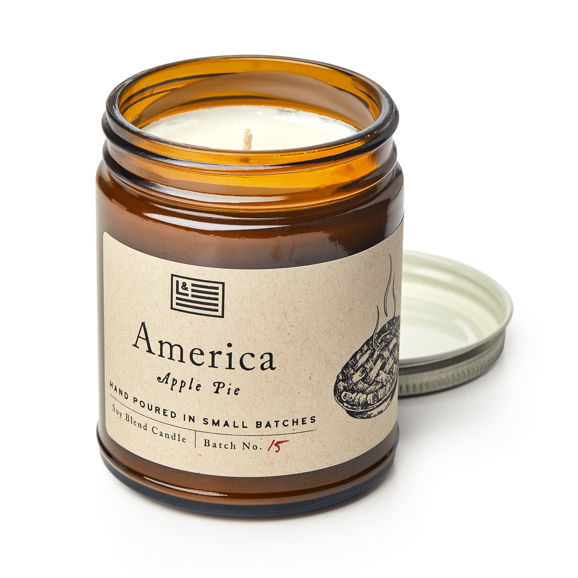 An America Candle in a brown glass jar, labeled "America Apple Pie" with a pie drawing and flag icon, sits with its metal lid. The partially used white soy blend candle creates a cozy atmosphere.