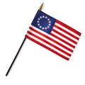 The Betsy Ross Stick Flag features 13 stars in a circle on a blue field and 13 red and white stripes, symbolizing early American independence. It is attached to a black stick with a gold spear tip.