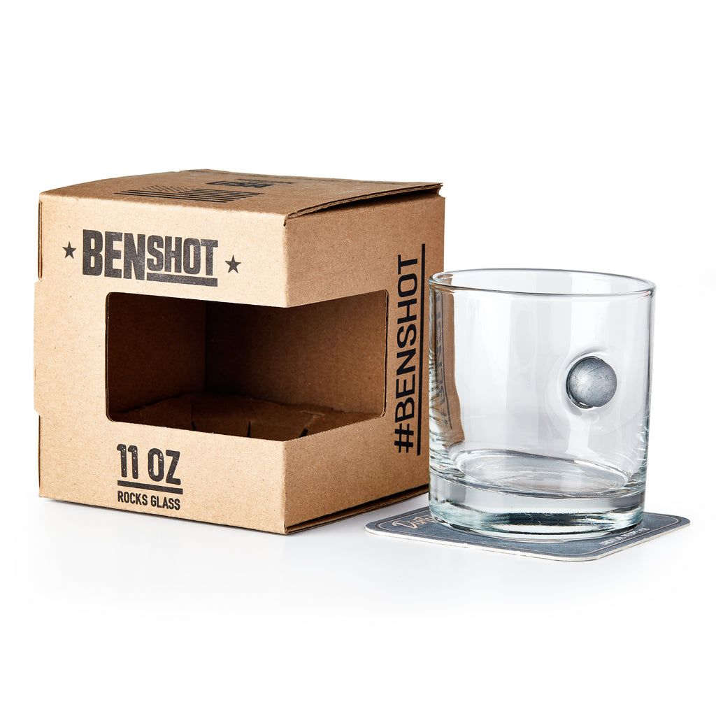The Musket Ball Rocks Glass is a clear, handcrafted 11 oz glass with a real musket ball embedded in its side. It sits on a square coaster beside BENSHOT packaging. Proudly made in the USA, this unique glass makes a bold statement.