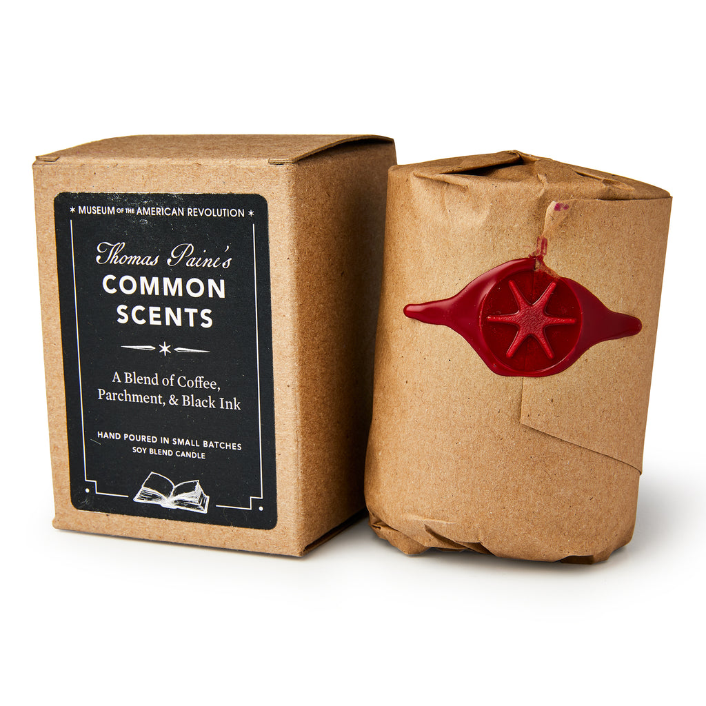 A Common Scents Candle in a brown box labeled “Thomas Paine’s Common Scents: A Blend of Coffee, Parchment, & Black Ink” stands beside a hand-poured soy blend candle wrapped in brown paper, sealed with red wax and a star emblem.