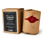 A Common Scents Candle in a brown box labeled “Thomas Paine’s Common Scents: A Blend of Coffee, Parchment, & Black Ink” stands beside a hand-poured soy blend candle wrapped in brown paper, sealed with red wax and a star emblem.