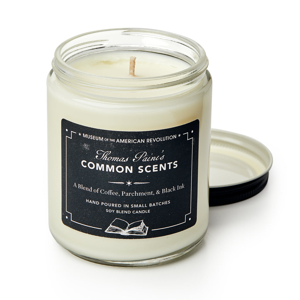 A glass jar soy blend "Common Scents Candle" with a black lid sits beside its label, which lists coffee, parchment, and black ink scents and features an open book illustration at the bottom.