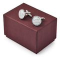 A pair of Declaration Cufflinks — round, silver, and engraved — rest on a closed maroon gift box.