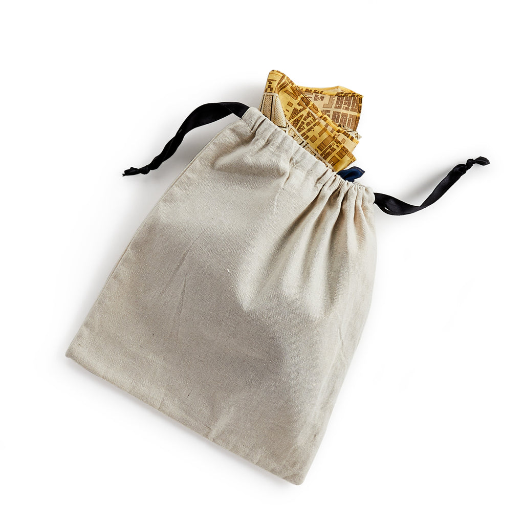 A beige fabric drawstring bag with black cords, partially open to reveal pieces of yellow patterned paper inspired by the Historic Philadelphia Then Scarf, featuring map designs, set against a white background.