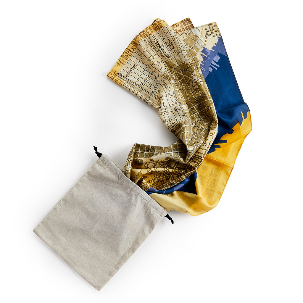 The Historic Philadelphia Then Scarf, featuring a city map design in yellow, blue, and brown, is partially pulled out of a beige drawstring pouch.