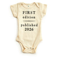 The First Edition 2026 Onesie is a cream-colored organic cotton baby outfit with black "FIRST edition published 2026" text, short sleeves, and bottom snaps—an eco-friendly and charming vintage-inspired baby gift.