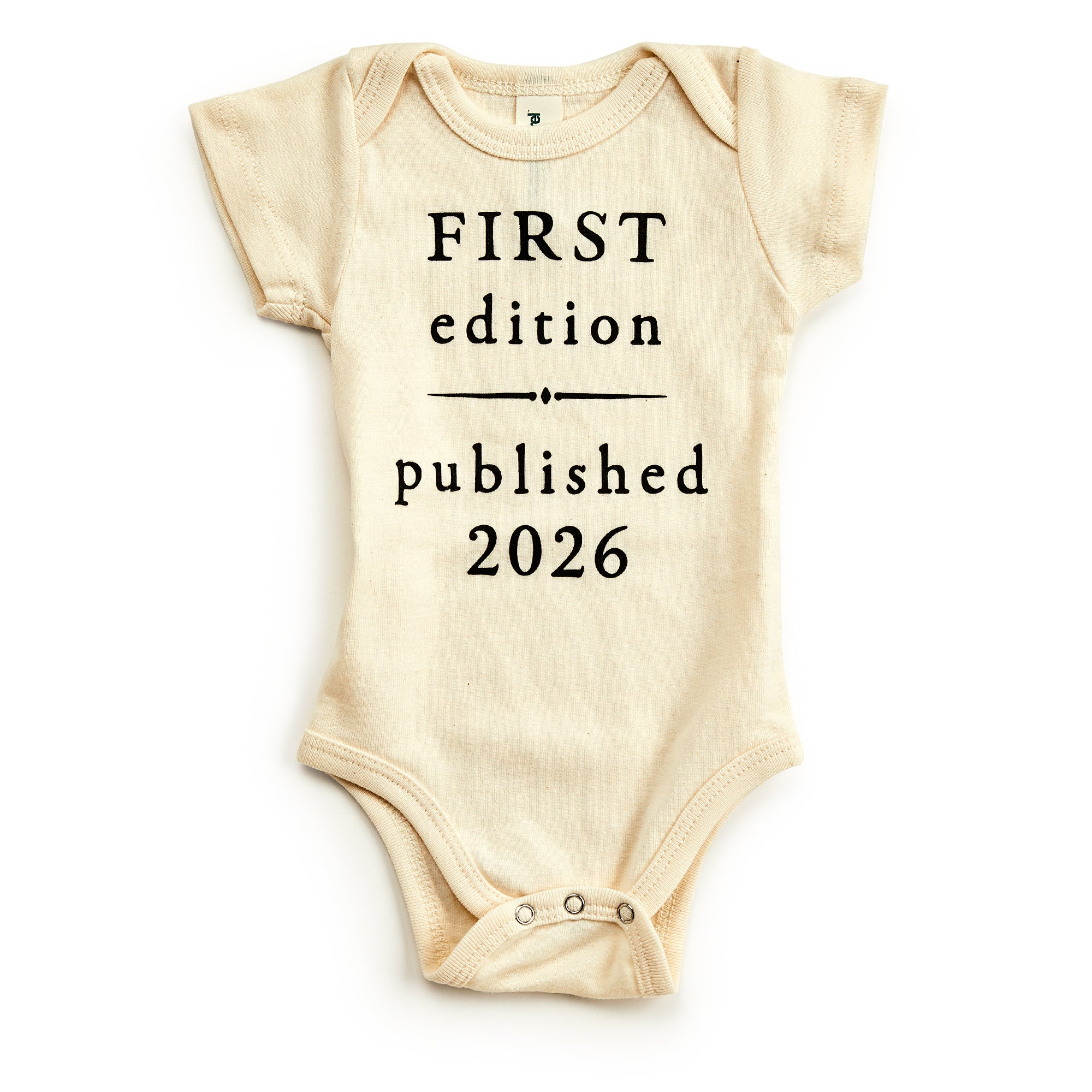 The First Edition 2026 Onesie is a cream-colored organic cotton baby outfit with black "FIRST edition published 2026" text, short sleeves, and bottom snaps—an eco-friendly and charming vintage-inspired baby gift.