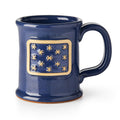 The George Washington Standard Mug is a dark blue stoneware mug with a rectangular tan patch displaying 13 raised star-like shapes inspired by the George Washington flag. It features a large rounded handle and slightly flared rim.
