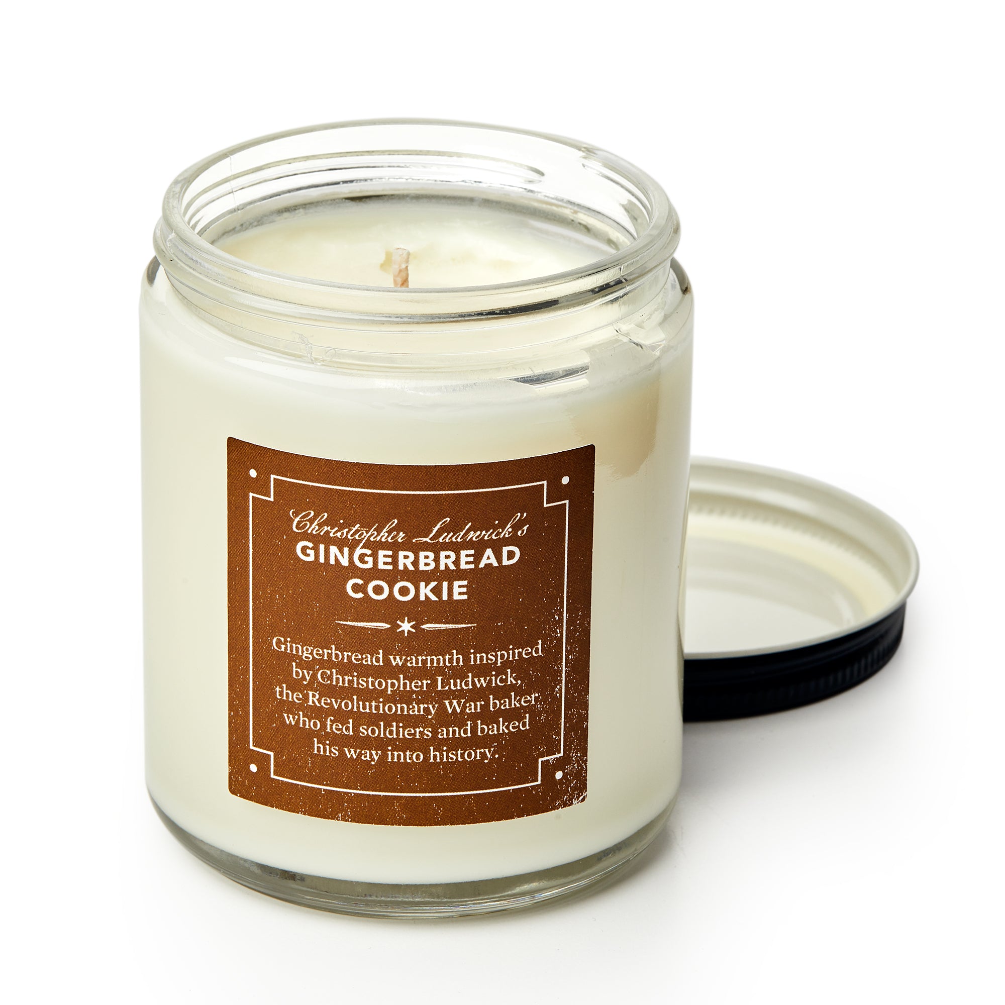 Christopher Ludwick's Gingerbread Candle features an open glass jar with a brown label, inspired by Revolutionary War baker Christopher Ludwick; the black lid rests beside it, filling your home with festive gingerbread fragrance.