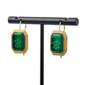 Glass Intaglio Earrings, Dark Green: Rectangular green glass earrings with gold frames and carved designs hang from gold-plated earwires on a black stand, recalling the refined elegance of colonial-era jewelry.
