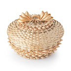 The Woven Basket is a round bamboo container with a lid, featuring indigenous craftsmanship and intricate ribbon bows and edging in light tan, displayed against a white background.