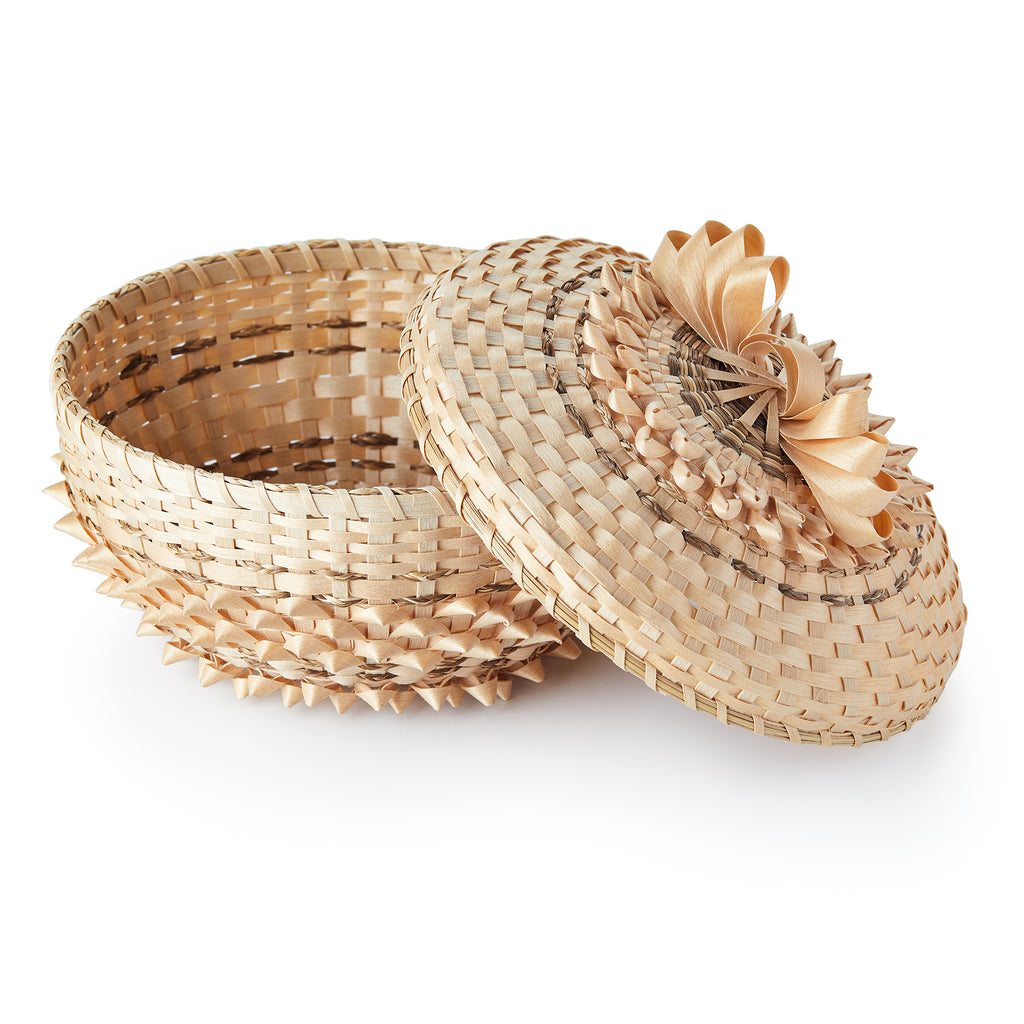 The Woven Basket, set against a white background, showcases Indigenous craftsmanship with a partly open round form and a decorative lid featuring an intricate light-colored floral pattern matching the basket’s woven design.