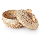 The Woven Basket, set against a white background, showcases Indigenous craftsmanship with a partly open round form and a decorative lid featuring an intricate light-colored floral pattern matching the basket’s woven design.