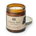 An unused Liberty Tree Candle in an amber glass jar labeled "Elm & Earth" features a beige tag with a tree and flag. Its metal lid rests nearby, ready to release the fresh elm and earth scent from its soy blend wax.