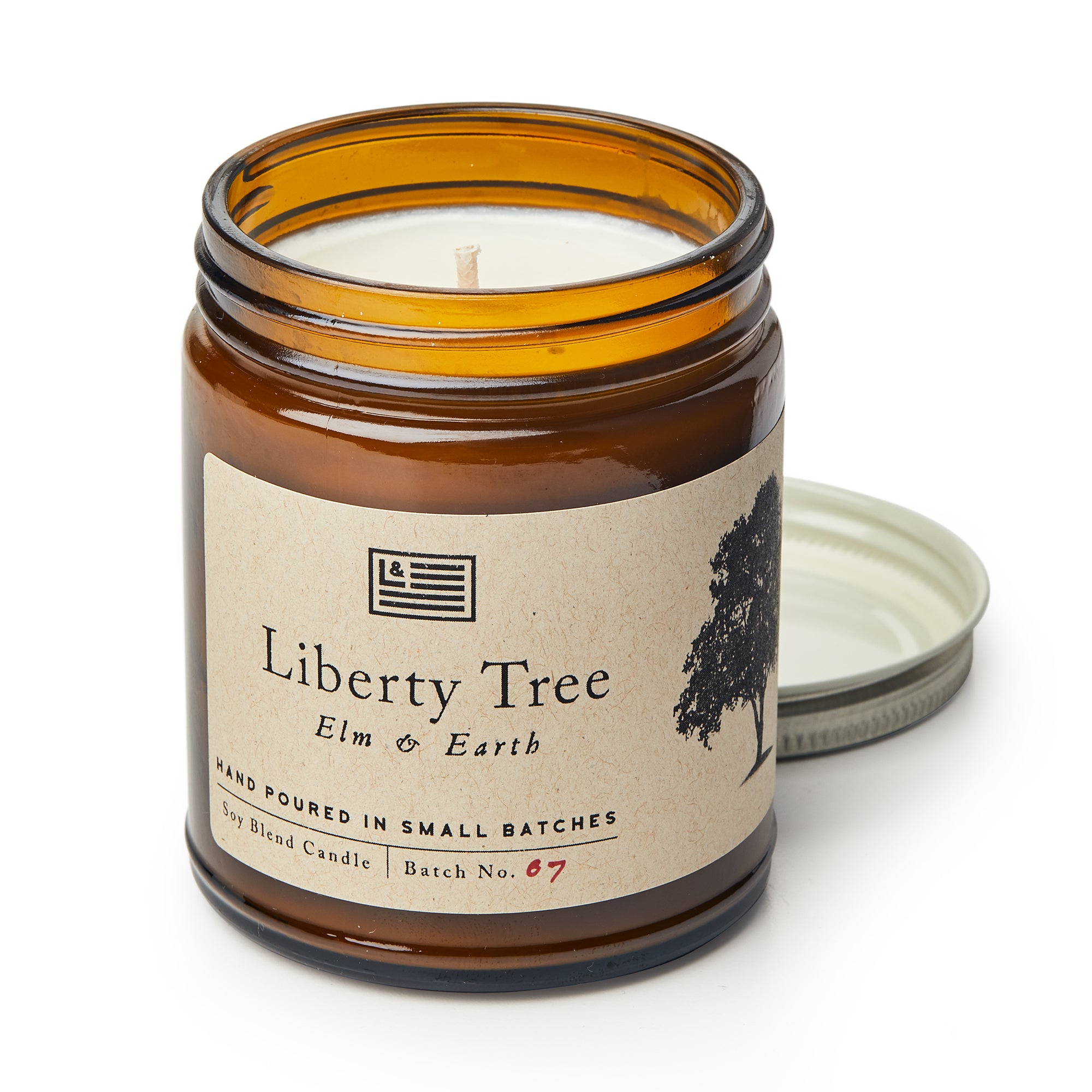 An unused Liberty Tree Candle in an amber glass jar labeled "Elm & Earth" features a beige tag with a tree and flag. Its metal lid rests nearby, ready to release the fresh elm and earth scent from its soy blend wax.