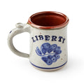 The Liberty Stoneware Mug is handcrafted with a brown interior, speckled beige exterior, blue floral design, a blue ring near the base, and "LIBERTY" on the side.
