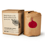 The Molly Pitcher Candle is a hand-poured soy candle wrapped in brown paper with a red wax seal, packaged in a box labeled “Molly Pitcher at the Battle of Monmouth—Quenching Relief” and adorned with a cannon to honor this heroine.