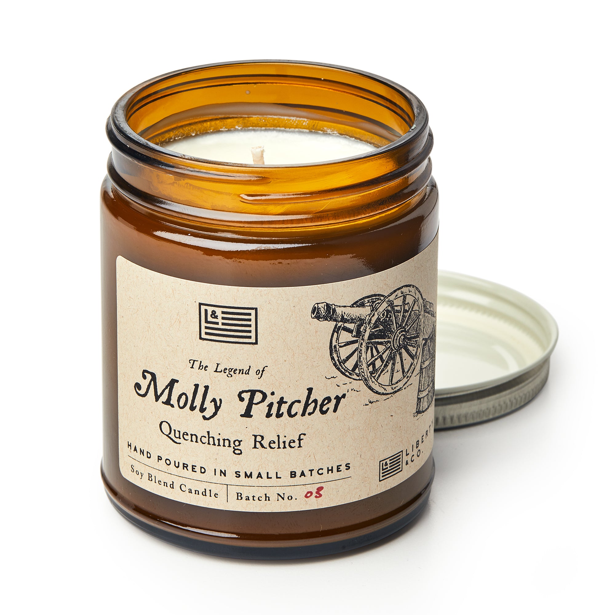 The Molly Pitcher Candle in an amber glass jar, labeled Quenching Relief, features a vintage cannon illustration. Its metal lid rests nearby, ready to unleash the citrus-scented soy aroma of this American Revolution-themed candle.