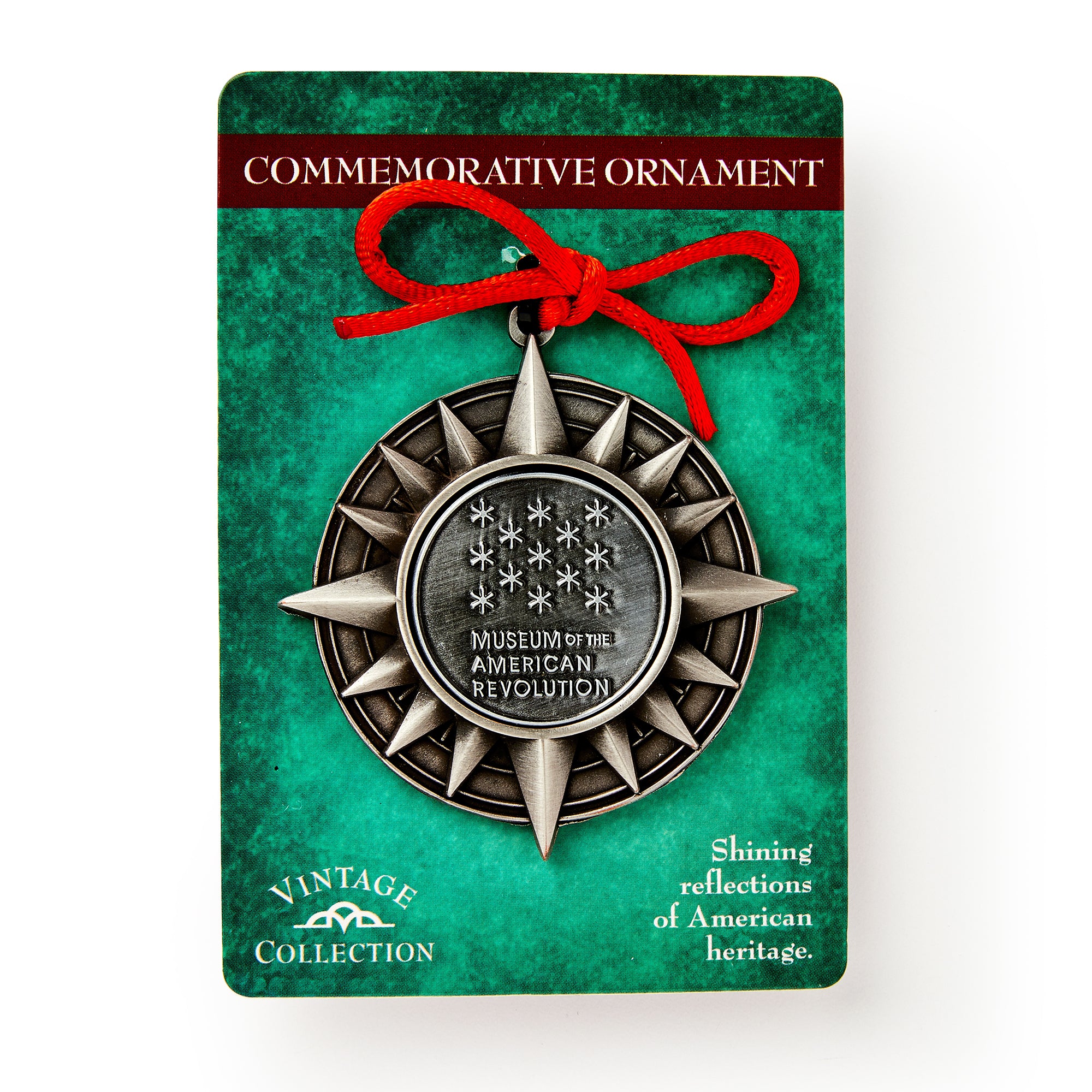 A pewter star-shaped ornament with a red ribbon, labeled Museum of the American Revolution, is displayed on green packaging that reads "Shining reflections of American heritage.