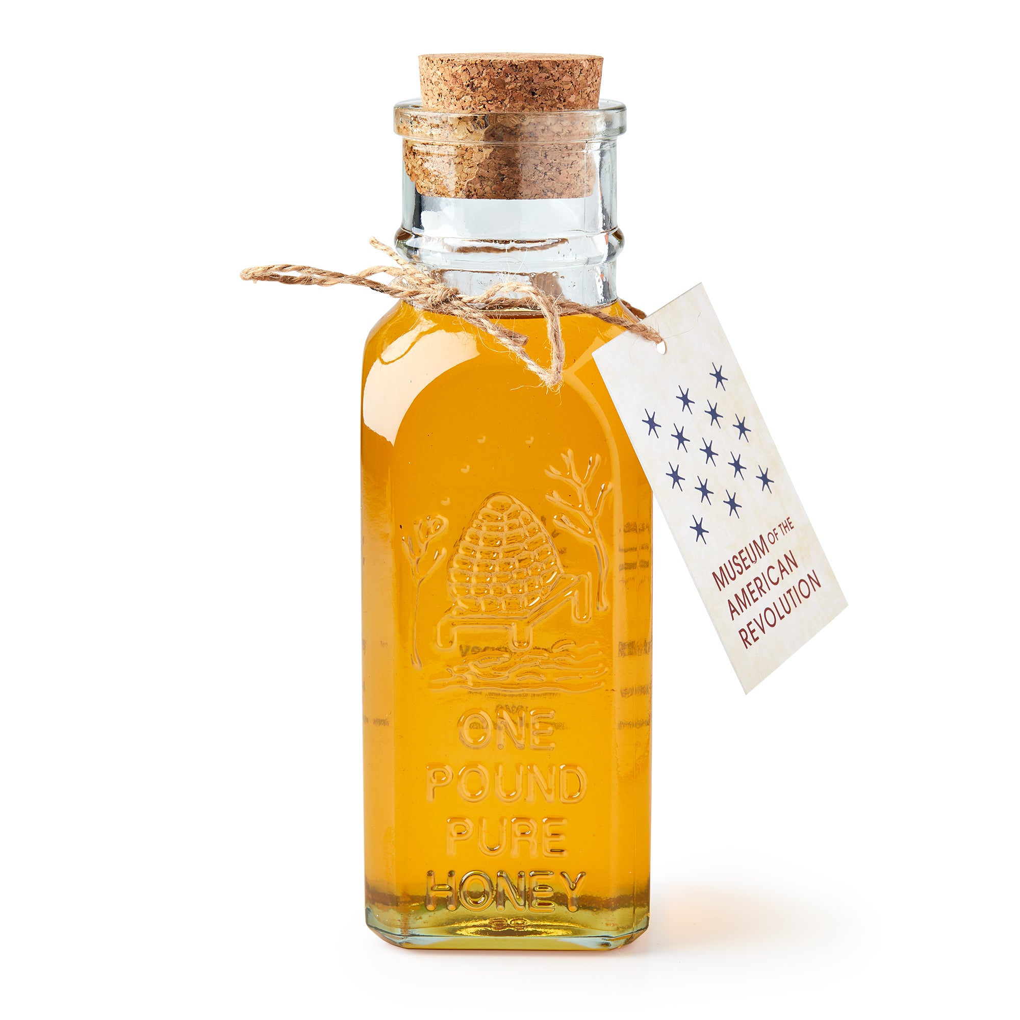 A clear glass bottle of Wildflower Honey, sealed with a cork and twine tag reading “Museum of the American Revolution,” features an embossed beehive design and the words “One Pound Pure Honey.”.