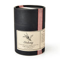 A black cylindrical container of Oolong Loose Leaf Tea features a beige label with a tea leaf illustration and is wrapped with a red-striped ribbon.